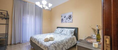 A bedroom with a bed, a chandelier, a painting, a bedside table with a bottle, and a window with curtains.