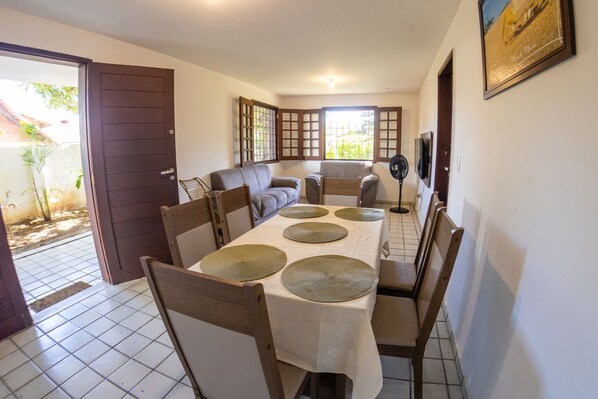 A dining area with a table set for four, surrounded by chairs, a sofa, and a fan. - Casa c Churrasqueira a 140m da Praia da Concei o (Paulista)