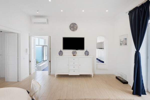 A modern hotel room with a large flat-screen TV, a white dresser, a window with a view, and a dark curtain. - Apartment 'Blue Room' with Sea View, Wi-Fi and Air Conditioning (Meta)