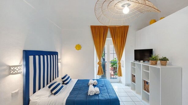 A hotel room with a bed, a TV, a shelf with plants, and a window with curtains. - Apartment 'Yellow Room' with Sea View, Wi-Fi and Air Conditioning (Meta)