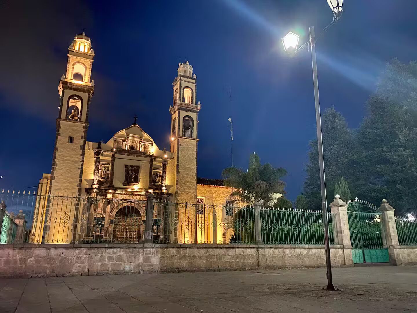 A historic building with two towers and a central dome, surrounded by a fence and a street lamp.