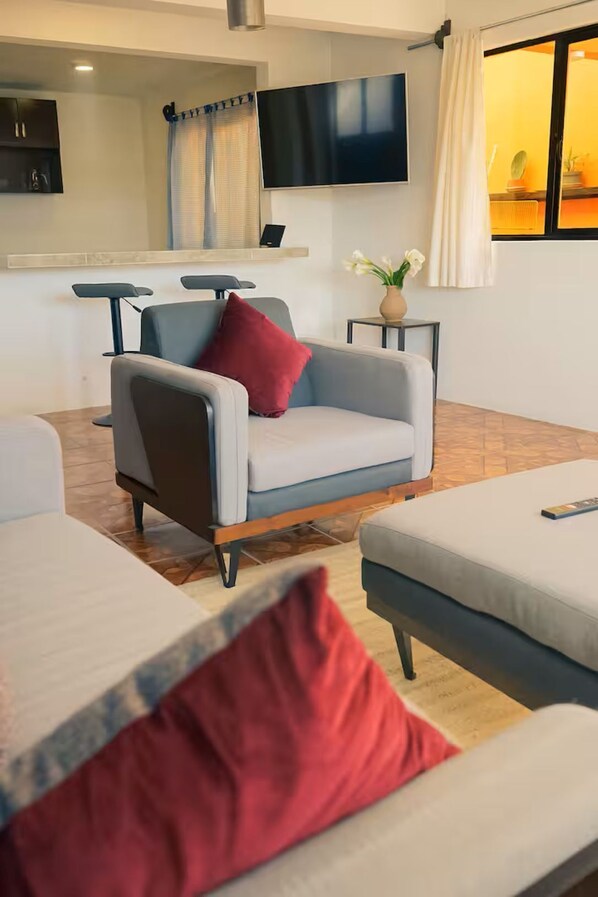 Un salon moderne avec un canapé gris, un coussin rouge, un téléviseur à écran plat et une petite table sur laquelle est posé un vase de fleurs.
