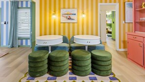 A room with striped walls, a beach-themed painting, two round tables, four green ottomans, and a pink cabinet. - Suncourt Retreat | Sports Court | Pool | Hot Tub (Bradenton)