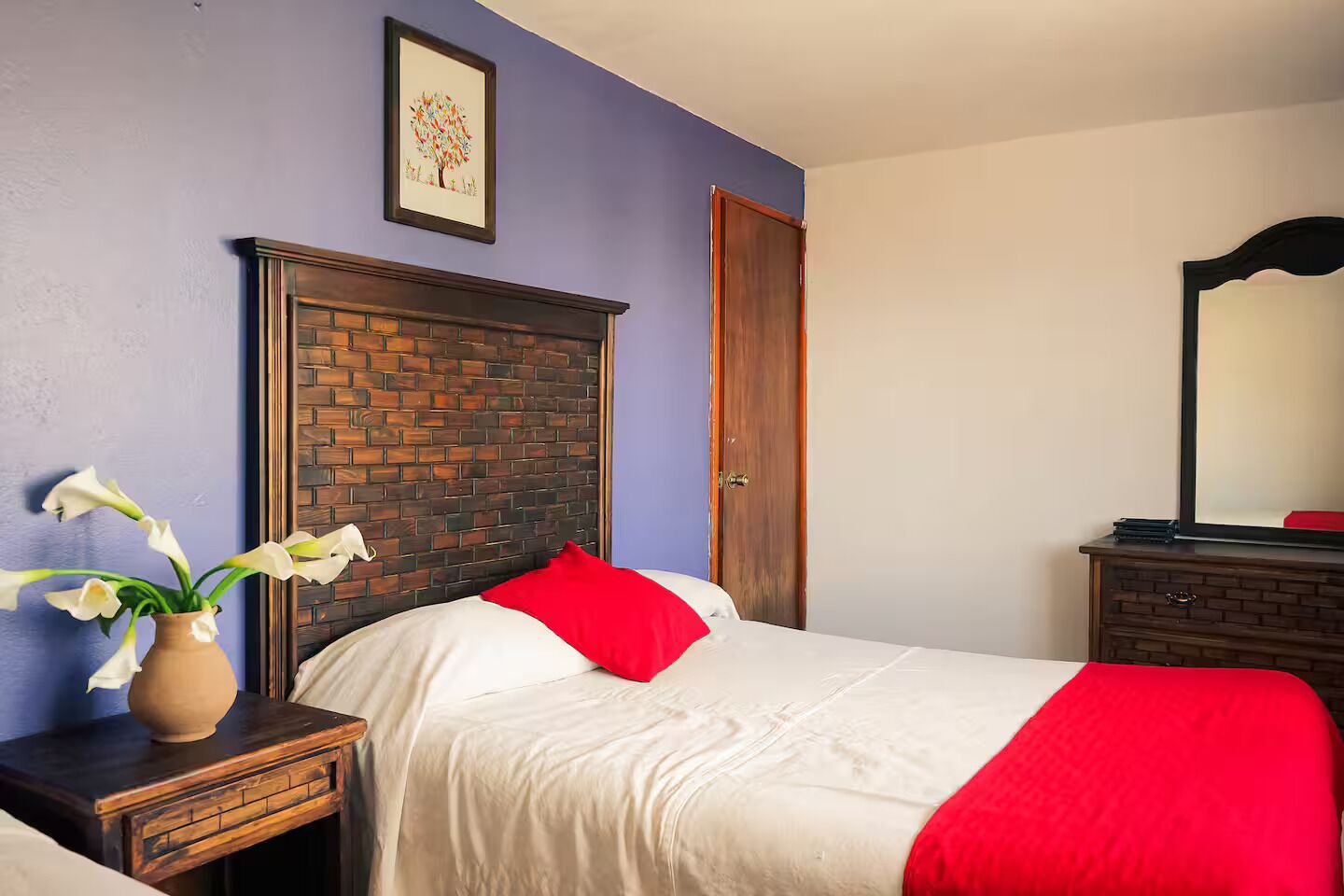 A bedroom with a brick-patterned headboard, a bed with white bedding and red pillows, a vase with white flowers on a nightstand, and a mirror on a dresser.