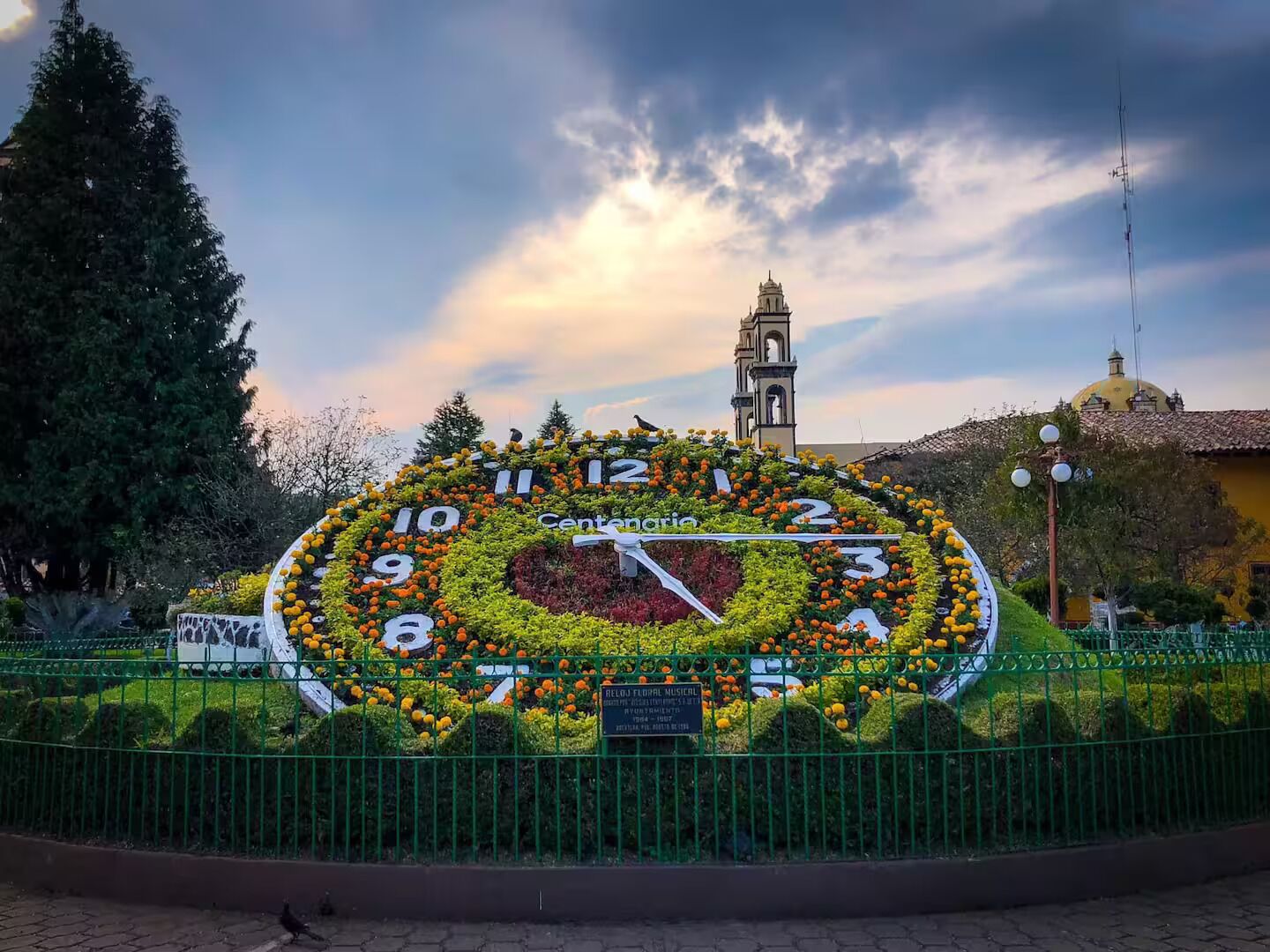 Eine große Außenuhr, umgeben von Blumen, mit einem Glockenturm und einem Gebäude im Hintergrund.