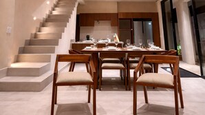 A modern dining area with a staircase leading to an upper level, a kitchen in the background, and a large window. - Jungle Villa + Private Studio w/ Rooftop Access | 2BR + Studio | Sleeps 6 (Tulum)
