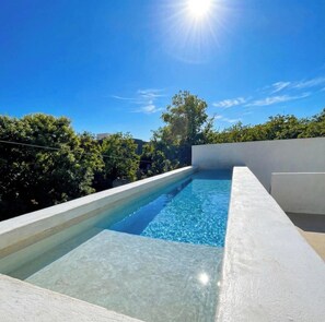 Une piscine d’un bleu éclatant, bordée de blanc, entourée de verdure et surplombée d’un ciel dégagé.