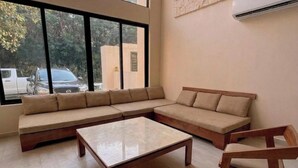 A modern living room with a sectional sofa, two wooden coffee tables, and a large window with a view of parked cars and trees. - Jungle Villa + Private Studio w/ Rooftop Access | 2BR + Studio | Sleeps 6 (Tulum)