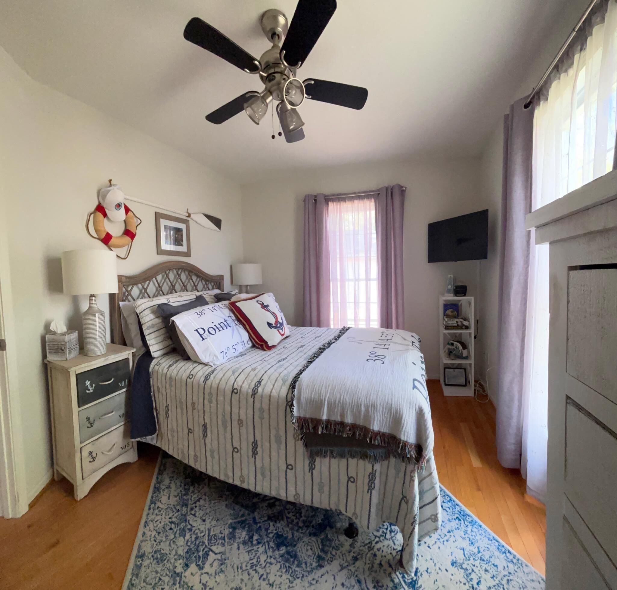 A bedroom with a bed, a ceiling fan, a TV, a nightstand, a lamp, a window with curtains, and a dresser.