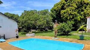 Ein rechteckiger Swimmingpool mit glasklar blauem Wasser, umgeben von einer Betonplattform. Im Hintergrund sind Kübelpflanzen und ein kleines weißes Gebäude zu sehen.