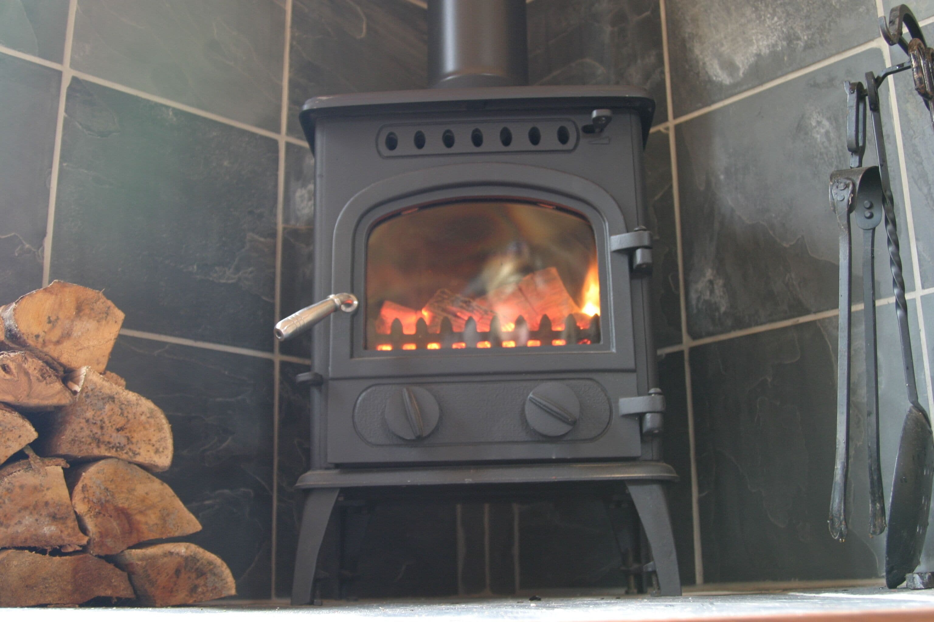 Un poêle à bois dans lequel le feu crépite, une pile de bois de chauffage à gauche et une poignée de porte en métal sur le poêle.