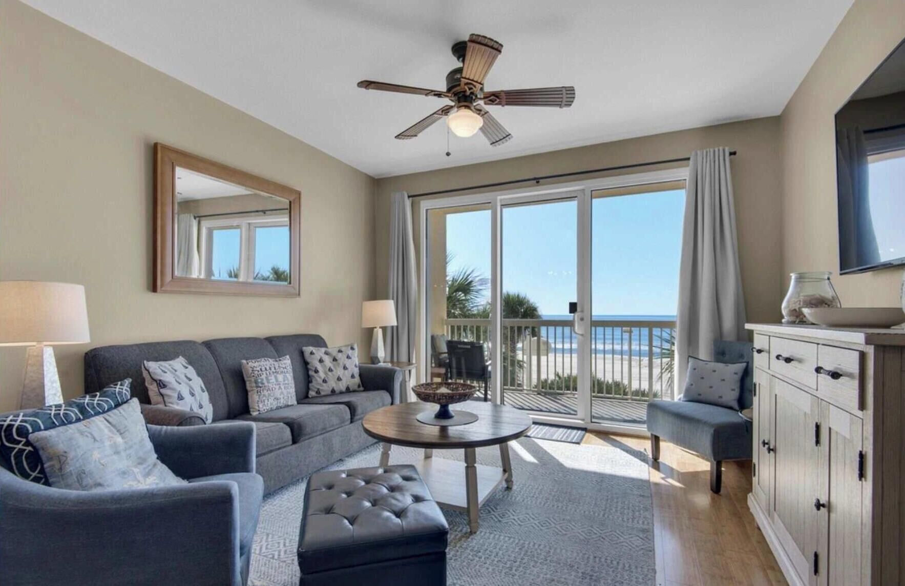 A living room with a sofa, ottoman, and a table, a ceiling fan, a large window with a view of the beach, and a cabinet.