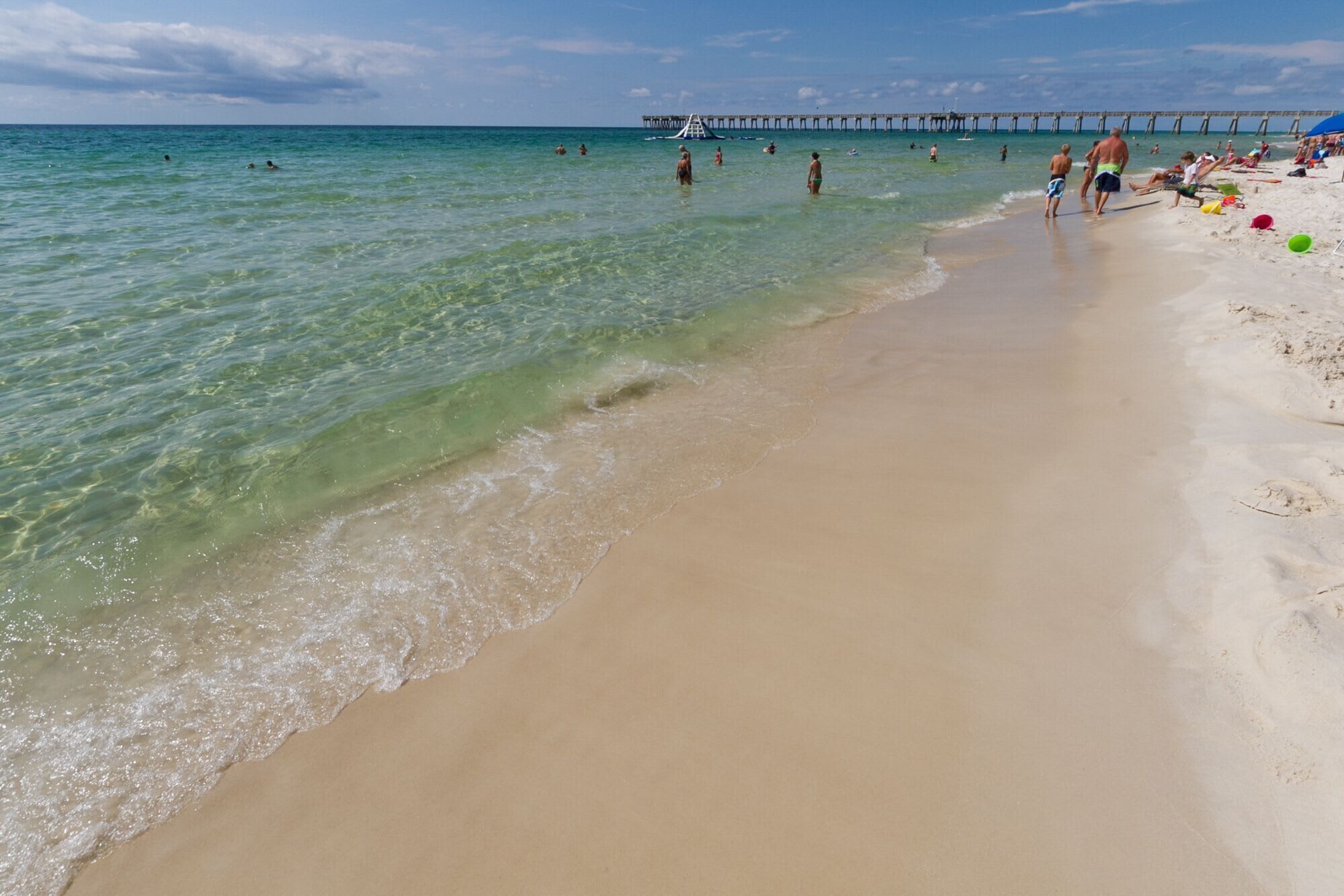 A beach with clear water, a pier in the background, and people on the sand.