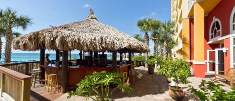A beachside bar with a thatched roof, outdoor seating, and a view of the ocean.