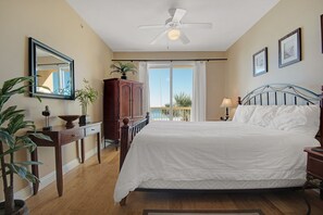 Une chambre à coucher comprenant un lit, un ventilateur de plafond, une fenêtre avec des rideaux, une table de chevet, une lampe, une commode et un miroir.