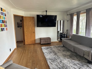 Ein Wohnzimmer mit Holzboden, einem grauen Sofa, einem Fernseher, einem Fenster mit Vorhängen und einer kleinen Korbschaukel.