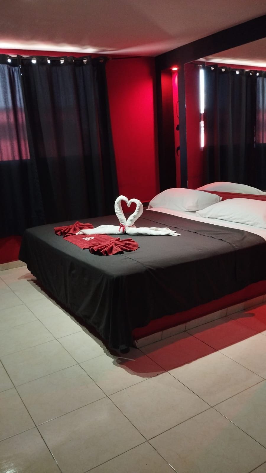 A hotel room with a bed, a heart-shaped towel arrangement, and red curtains.