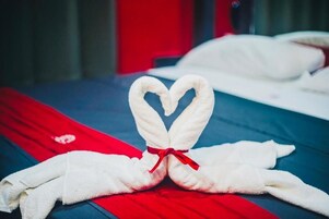 Ein Bett mit einer roten und blauen Tagesdecke, ein weißes Handtuch sowie eine herzförmige Handtuchdeko mit roter Schleife.