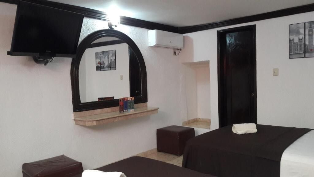 Una habitación de hotel con cama, televisor montado en la pared, espejo, estante con objetos y una puerta.