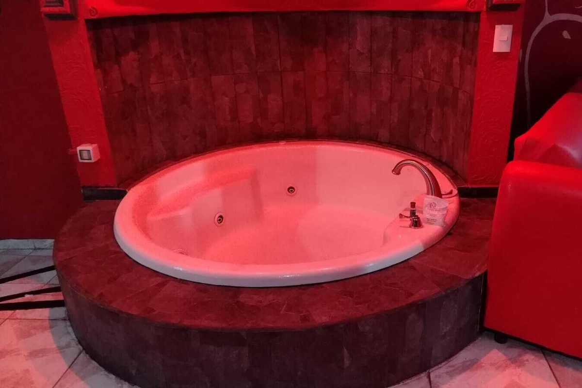 Una bañera redonda con un grifo, sobre una plataforma circular con base roja, en una habitación con paredes rojas y un panel de madera al fondo.