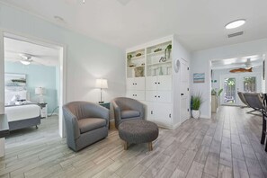 A room with a bed, two armchairs, a side table, a lamp, a cabinet with shelves, and a door leading to another room. - Sandy Toes Family Retreat (Jacksonville Beach)