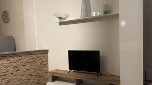 A modern living room with a flat-screen TV on a wooden stand, a stone accent wall, a white cabinet, and a shelf with decorative items.