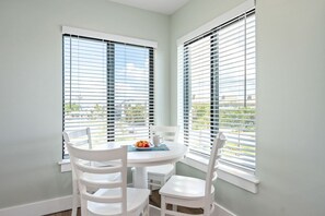 Un área de comedor con una mesa redonda y sillas blancas, ventanas amplias con persianas y vistas a zonas verdes.