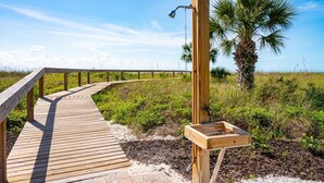 Une promenade en bois mène à une structure également en bois, où se trouvent une fontaine et, en arrière-plan, un palmier.