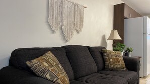 A dark grey sofa with patterned cushions, a macrame wall hanging, a lamp, and a refrigerator in a kitchen area. - Tranquil Candle Lake Townhouse Backing the 5th Green (Candle Lake)