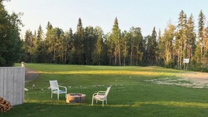A grassy outdoor area with two white chairs, a brick fire pit, and a wooden fence. - Tranquil Candle Lake Townhouse Backing the 5th Green (Candle Lake)