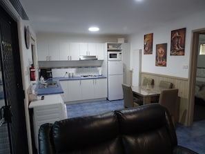 Une cuisine compacte avec des meubles blancs, un four à micro-ondes et une cuisinière à gaz. On y trouve un coin repas avec une table et des chaises, ainsi qu’un espace de vie avec un canapé en cuir.
