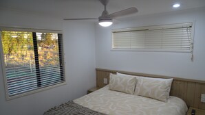 Un dormitorio con cama, ventilador de techo, ventana con persianas y mesita de noche.
