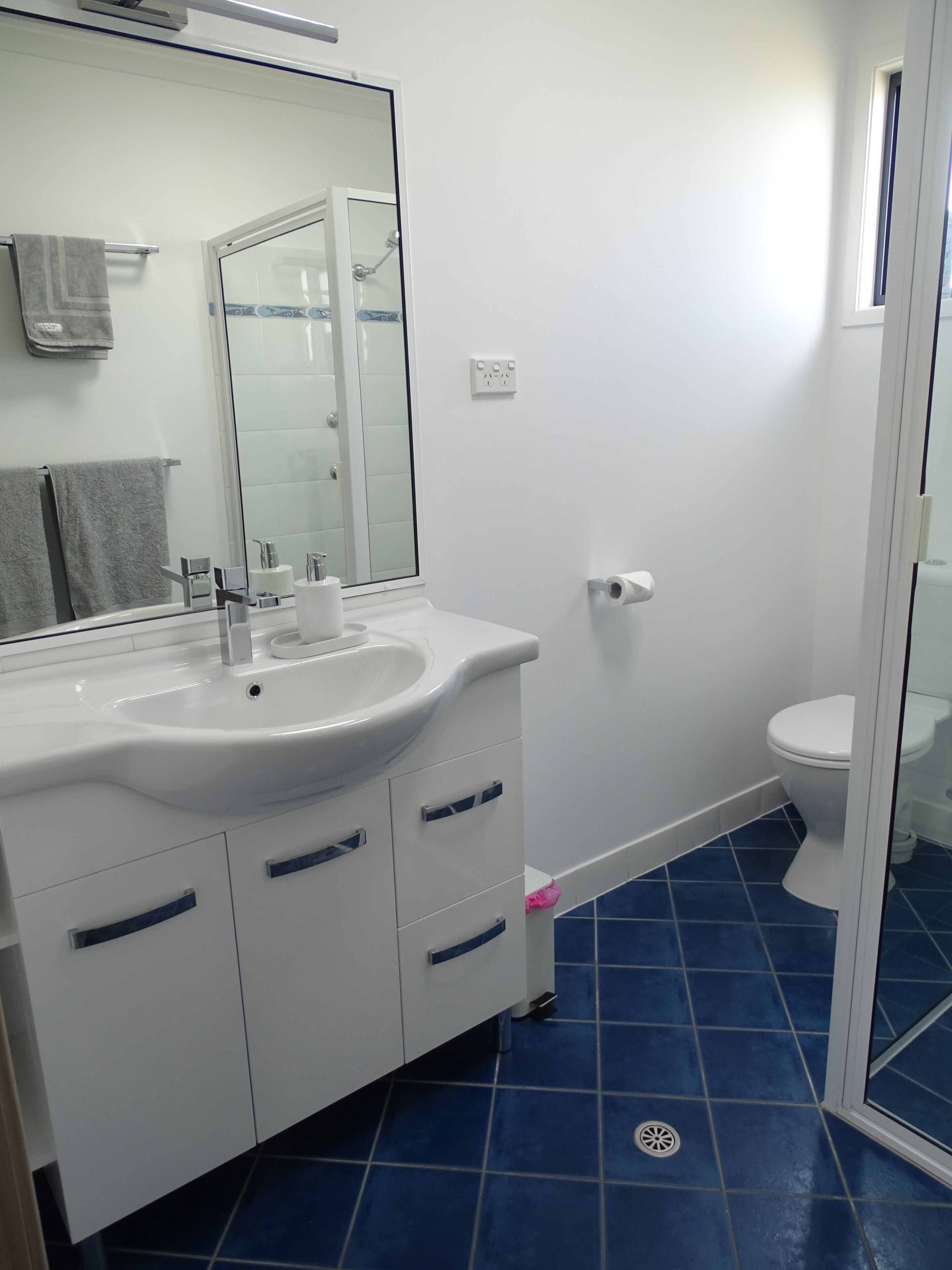 Ein Badezimmer mit weißem Waschbecken, Spiegel und Toilette, blau gefliestem Boden und einer Duschkabine aus Glas.