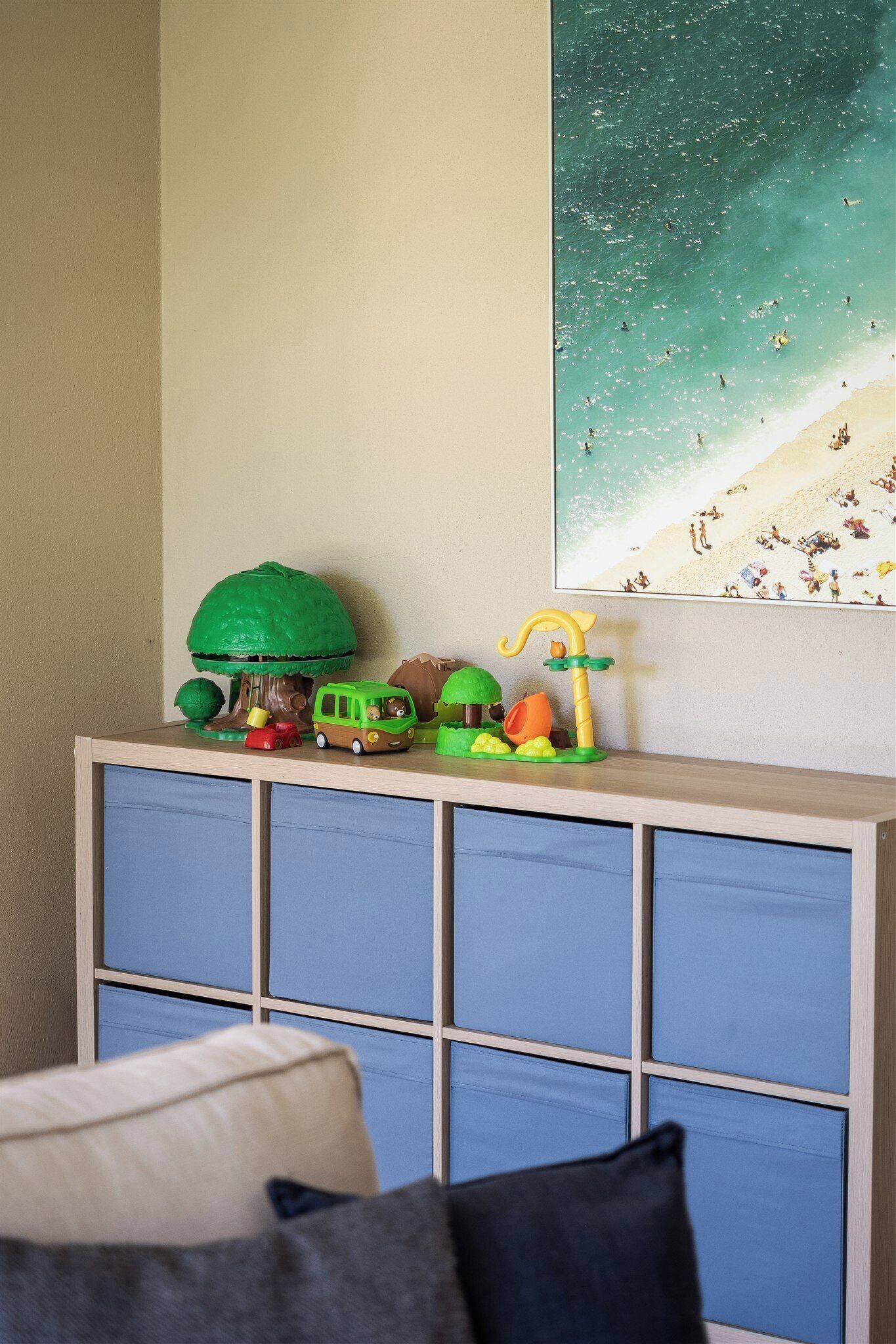 A room with a blue cabinet, a beach scene painting, and a toy setup including a green tree, a yellow toy car, and a brown toy vehicle.