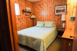 Et rum med træpaneler, en seng, et natbord, en lampe og et billede i ramme på væggen.