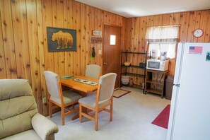 Ein Zimmer mit Holzwänden, einem Esstisch mit Stühlen, einem Kühlschrank, einer Mikrowelle und einem kleinen Fenster mit Vorhängen.