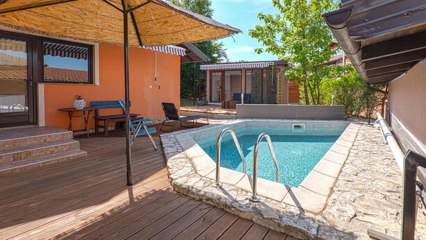 Piscina al aire libre con escalones de acceso, rodeada por una terraza de madera y un área de estar cubierta.