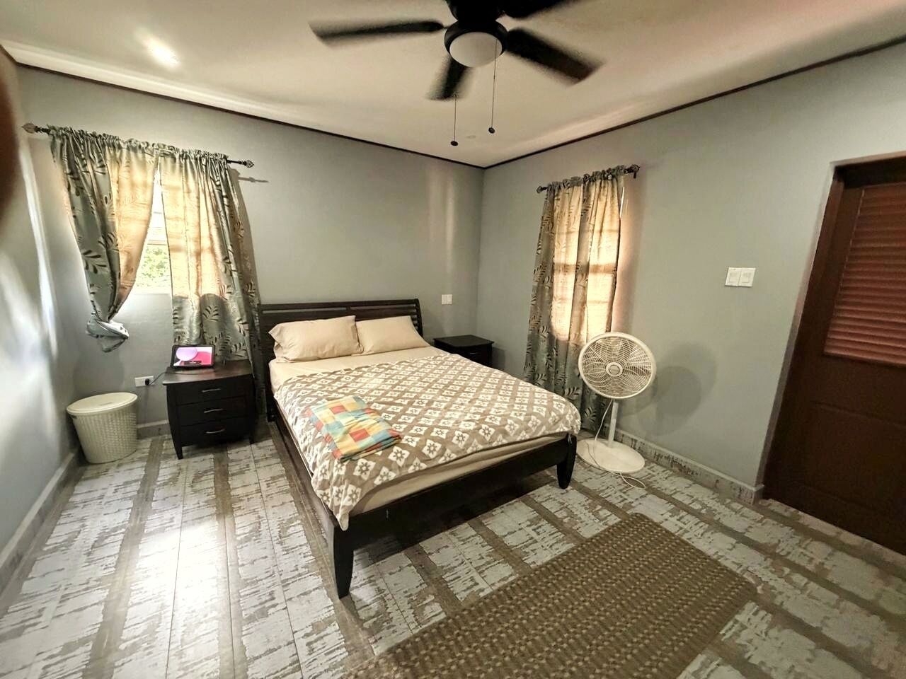 Ein Schlafzimmer mit Bett, Nachttischen, einem Ventilator, Deckenventilator und einem Fenster mit Vorhängen.