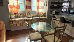 Un ambiente abitativo compatto con un tavolo da pranzo in vetro, sedute imbottite, angolo cottura e una finestra con tende.