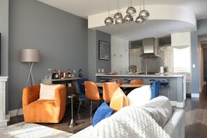 Ein modernes Wohnzimmer mit einer Couch, Sesseln, einem Couchtisch und einem Essbereich, im Hintergrund eine Küche.