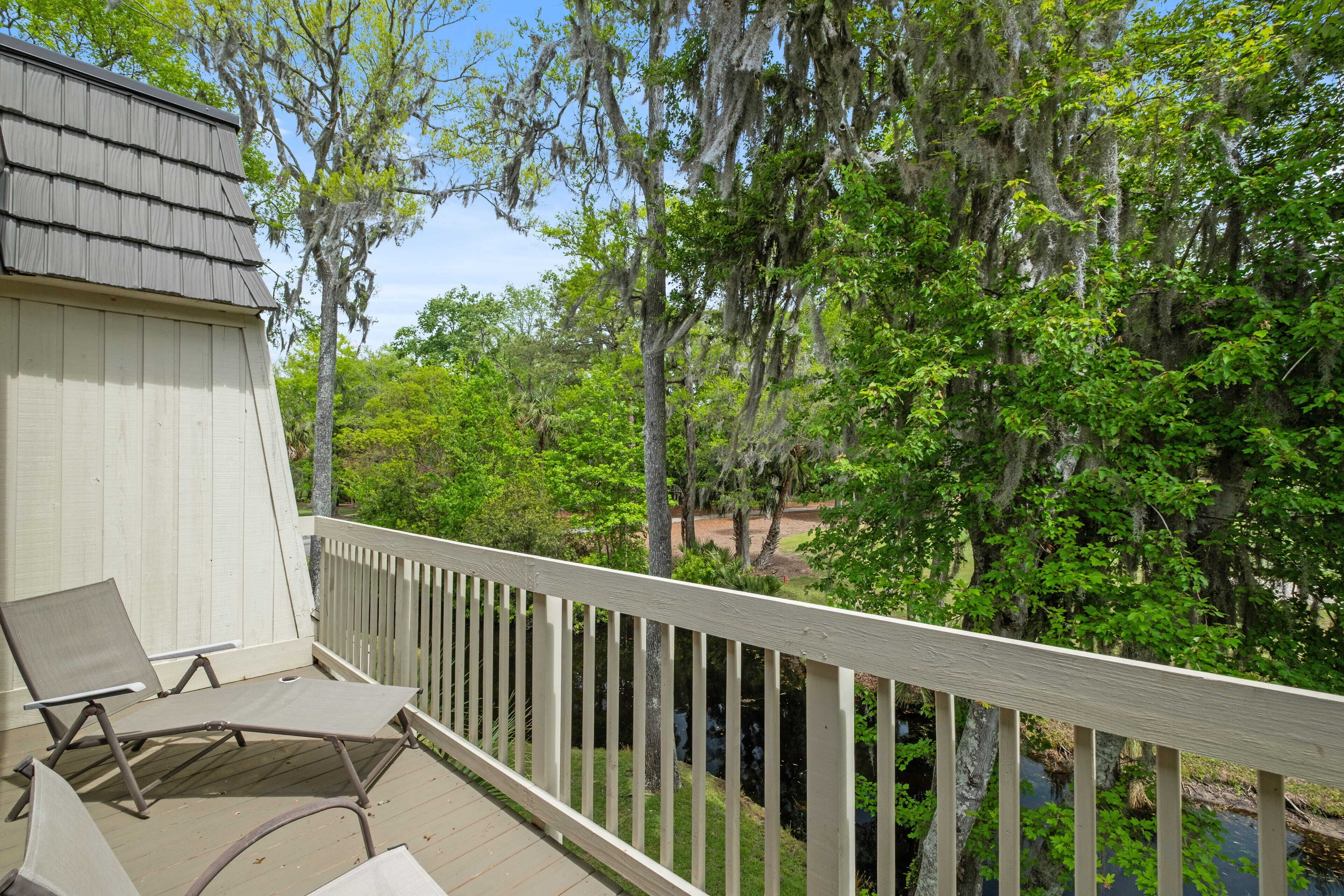 A wooden deck with two lounge chairs, a white railing, and a view of a wooded area with trees and a water body.