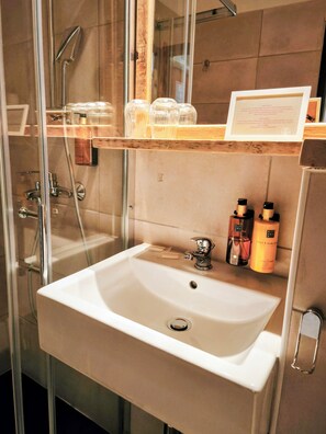 Un baño con ducha de vidrio, un lavabo blanco y un estante de madera con artículos de aseo.