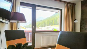 A room with a view of a mountainous landscape through a large window, a table, two chairs, a lamp, and a radiator.