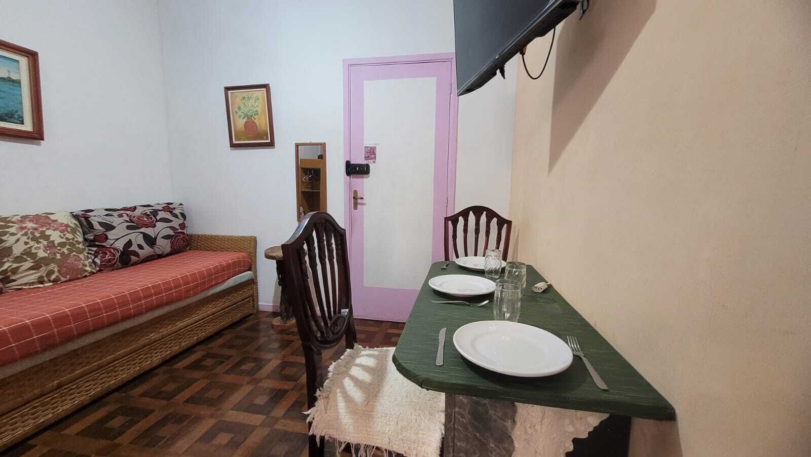 A small room with a dining table set for two, a sofa with floral cushions, a wooden chair, a pink door, and a wall-mounted TV.