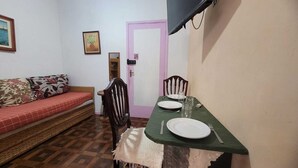 Una habitación pequeña con una mesa de comedor dispuesta para dos personas, un sofá con cojines florales, una silla de madera, una puerta rosa y un televisor montado en la pared.