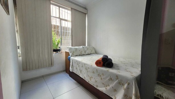 Un dormitorio pequeño con cama, una ventana con cortinas, una planta en maceta y suelo de baldosas.