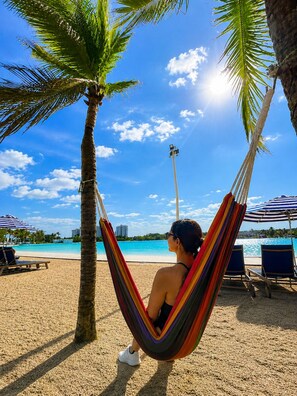 A hammock under a palm tree on a beach with a clear sky and a view of the ocean.