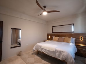 Un dormitorio con una cama, un ventilador de techo, un espejo montado en la pared y una mesita de noche.