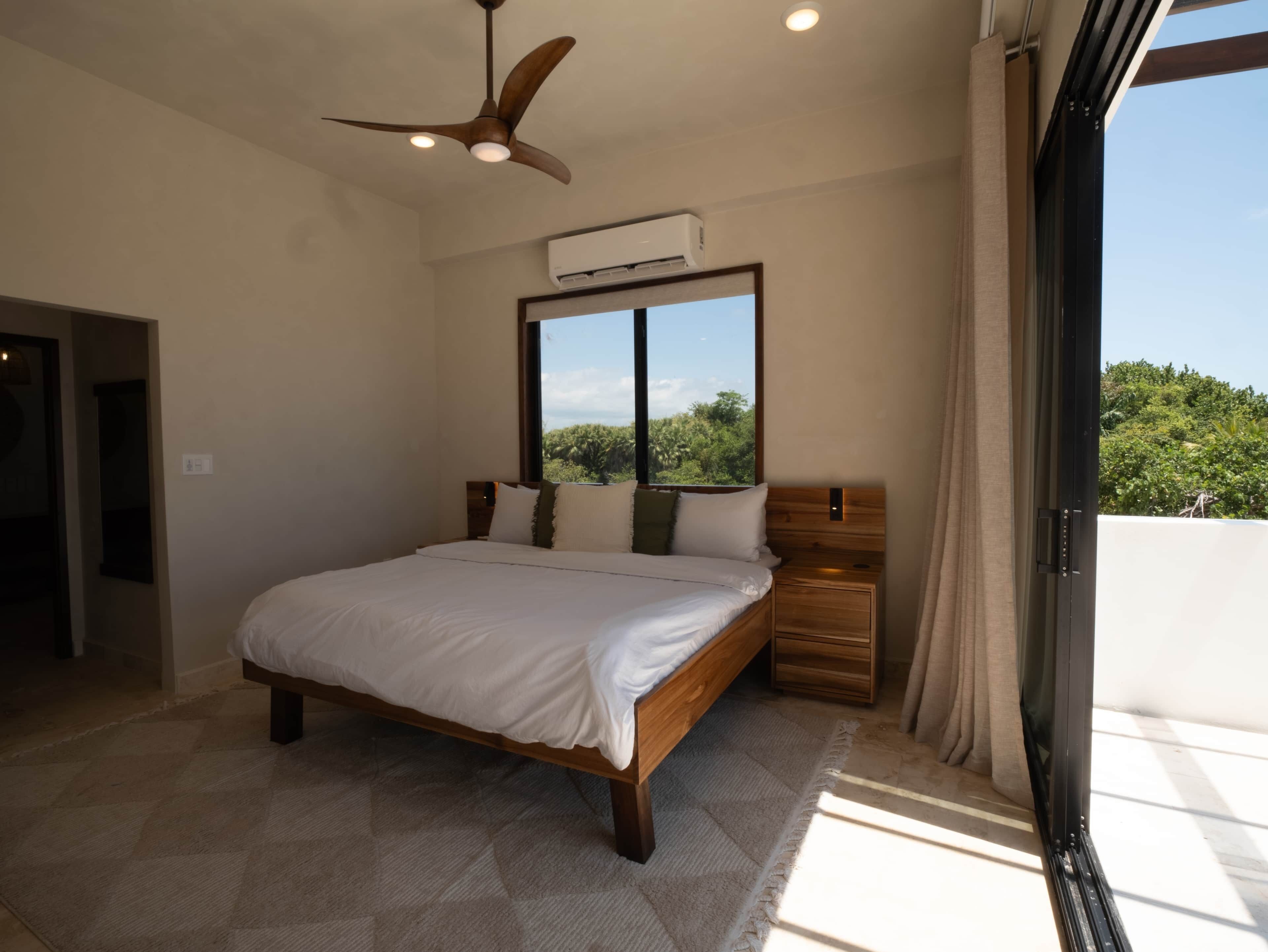 A bedroom with a bed, a ceiling fan, an air conditioning unit, a window with a view, and a door.