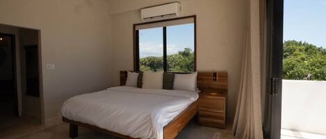 A bedroom with a bed, a ceiling fan, an air conditioning unit, a window with a view, and a door.